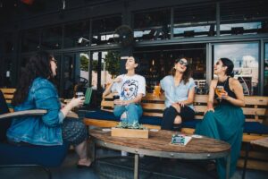 Photo of four women laughing while sitting outside on a bars terrace having a drink together.