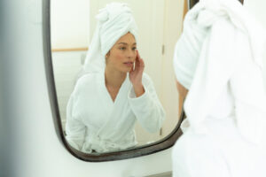 Photo of a woman in a bathrobe looking into a mirror and touching her face.