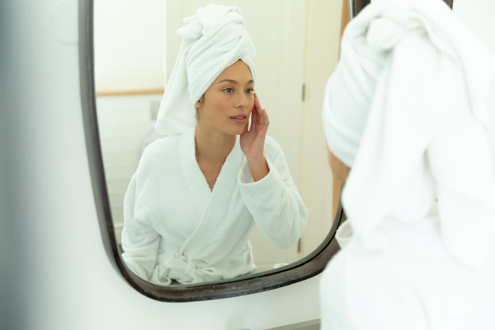 Photo of a woman in a bathrobe looking into a mirror and touching her face.